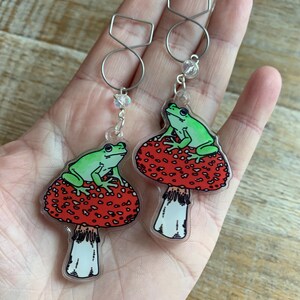 May include: A pair of earrings featuring a green frog sitting on a red and white mushroom. The earrings are made of clear acrylic and have silver-toned hooks.