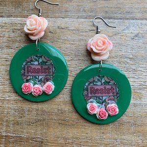 May include: A pair of green dangle earrings. Each earring features a pink rose at the top, a green circle with the word "Resist" in a floral frame, and three small pink roses at the bottom. The earrings hang from silver hooks.