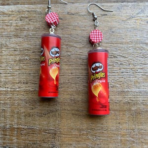 May include: A pair of novelty earrings featuring miniature Pringles cans. The red cans have the Pringles logo and are suspended from silver-colored fish hook ear wires. Each earring has a red and white checkered bead above the can.