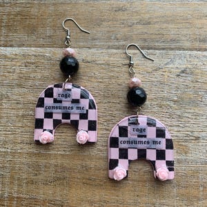 May include: A pair of handmade earrings with a pink and black checkered design. Each earring features a pink arch shape with the words "rage consumes me" and small pink rose accents. The earrings have silver hooks and black and pink bead accents.