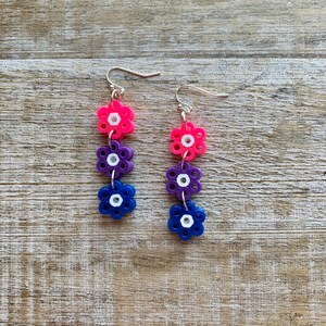 May include: A pair of dangle earrings with three flower-shaped beads on each earring. The beads are pink, purple, and blue with white centers.