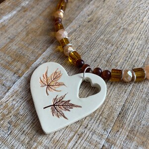May include: A white ceramic heart pendant necklace with a cutout heart and two brown leaf designs. The necklace is made with brown and clear glass beads.