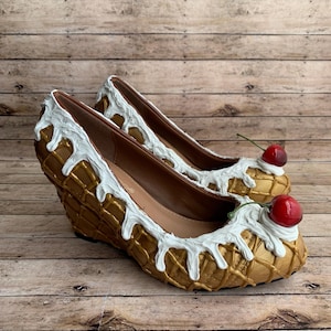 May include: A pair of novelty wedge heels styled as ice cream cones. The shoes have a gold waffle cone design, white frosting detail, and a red cherry. The heels are approximately 7.6 cm high.