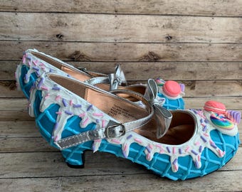 May include: A pair of whimsical blue Mary Jane style shoes with white icing and colorful sprinkles. The shoes feature silver straps and bows, and are decorated with macarons and candy.