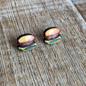 May include: A pair of wooden earrings shaped like hamburgers. The earrings are painted with realistic details, including a sesame seed bun, lettuce, tomato, cheese, and a meat patty.