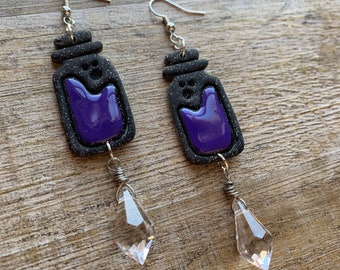 Peut inclure: Une paire de boucles d'oreilles pendantes avec des bocaux en verre noir remplis de liquide violet et des breloques en cristal transparent en forme de larme.