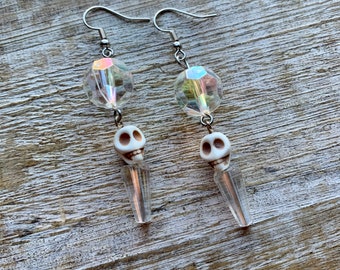 May include: A pair of silver-tone dangle earrings. Each earring features a clear, faceted bead, a small white skull charm, and a clear, rectangular charm. The earrings are displayed on a wooden surface.