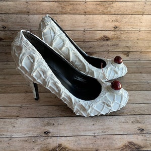 May include: White high-heeled shoes with a textured waffle cone design and red cherry accents.