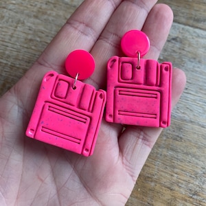 Puede incluir: Un par de pendientes de disquete rosa con textura moteada. Los pendientes están hechos de arcilla polimérica y tienen un acabado brillante.