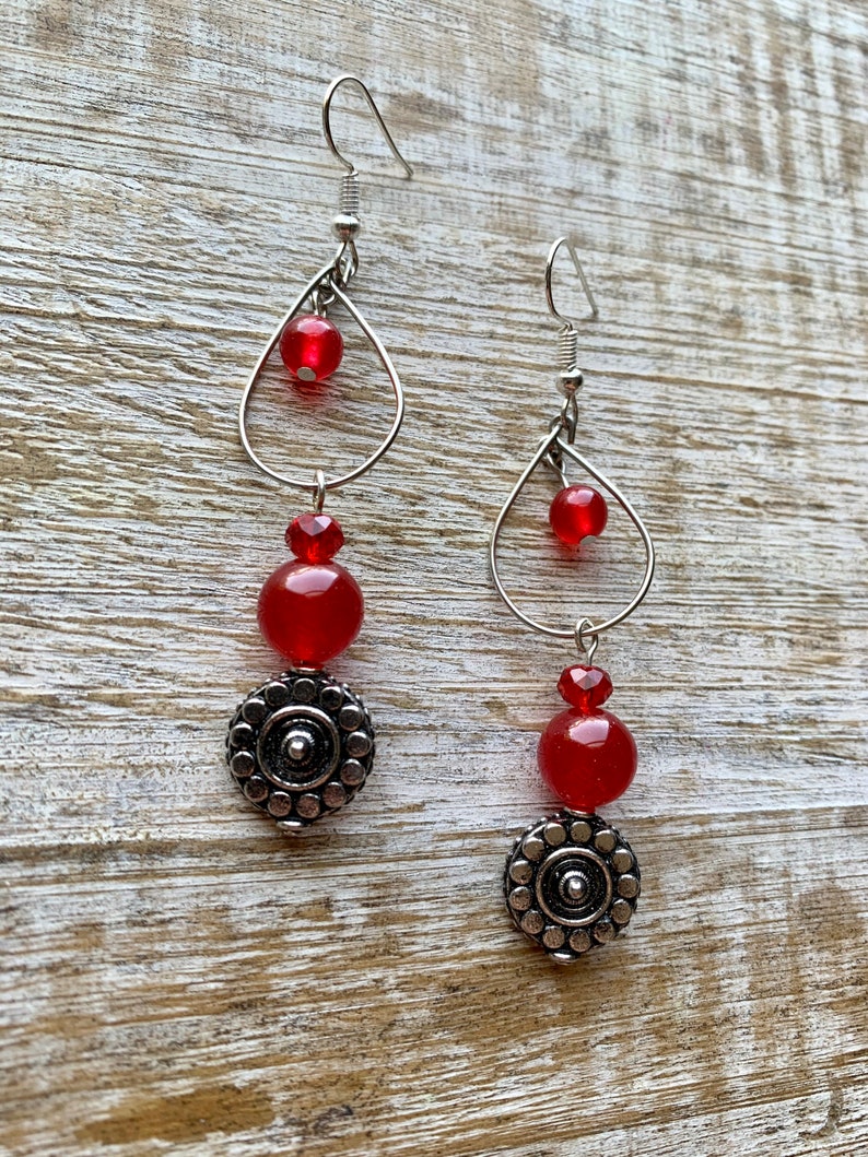 Op de afbeelding: Een paar oorhangers met rode kralen en zilveren accenten. De oorhangers hebben een traanvormig draadframe met een kleinere rode kraal bovenaan en een grotere rode kraal eronder. De onderkant van de oorhangers is versierd met een zilveren ronde hanger.