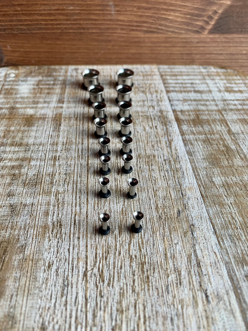 May include: A set of 12 silver ear gauges, arranged in size order from largest to smallest, on a wooden surface.