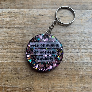 May include: A round keychain with a glittery, dark background. The keychain has the text "men of quality do not fear equality" in a white font. The keychain is attached to a silver metal ring.