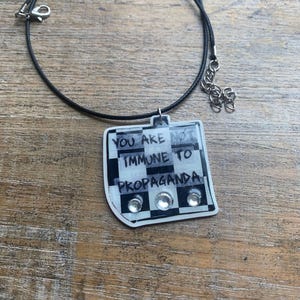 May include: A black cord necklace with a pendant that reads "YOU ARE NOT IMMUNE TO PROPAGANDA." The pendant has a black and white checkered pattern and three clear rhinestones.