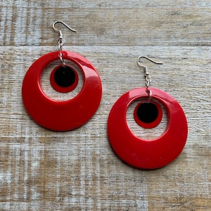 May include: A pair of red hoop earrings with a black circle cutout in the center. The earrings have silver hooks.