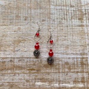 Op de afbeelding: Een paar oorhangers met rode glazen kralen en zilveren metalen accenten. De oorhangers hebben een traanvorm met een zilveren draadlus en een zilveren kraal aan de onderkant.