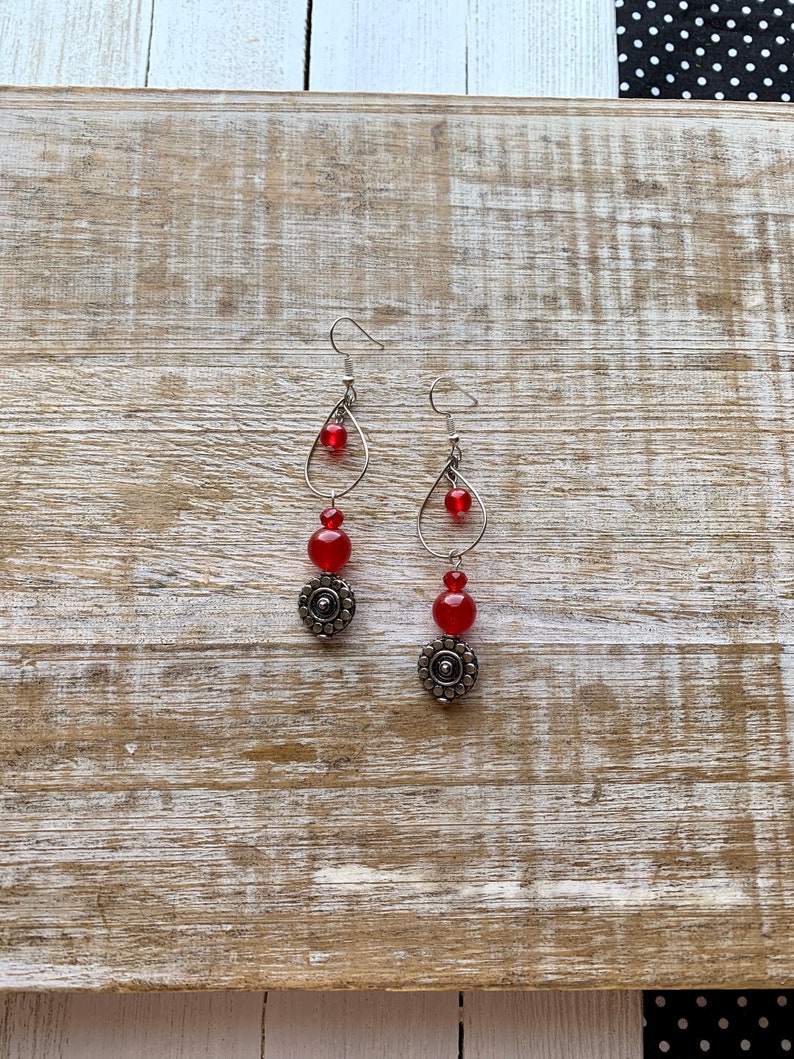 Op de afbeelding: Een paar oorhangers met rode glazen kralen en zilveren metalen accenten. De oorhangers hebben een traanvorm met een zilveren draadlus en een zilveren kraal aan de onderkant.