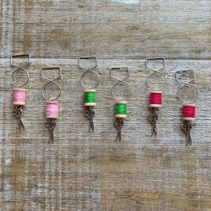 May include: Six metal wine bottle charms with a spool of thread and a pair of scissors hanging from each charm. The spools of thread are pink, green, green, red, and red.