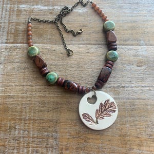 May include: A handcrafted necklace featuring a pendant with a leaf design and a heart-shaped cutout. The necklace has a mix of brown, green, and tan beads, and a bronze-colored chain.