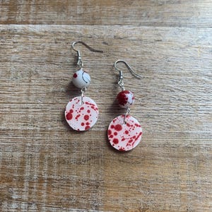 Puede incluir: Un par de pendientes colgantes con un patrón de salpicaduras blancas y rojas. Los pendientes tienen un gancho plateado y una pequeña cuenta blanca con salpicaduras rojas.