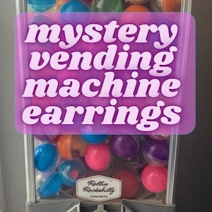 Può includere: Un distributore automatico blu e argento pieno di palline di plastica colorate. La macchina ha un cartello che dice "Rollin' Rockabilly" e una fessura per le monete. La macchina è etichettata "mystery vending machine earrings".