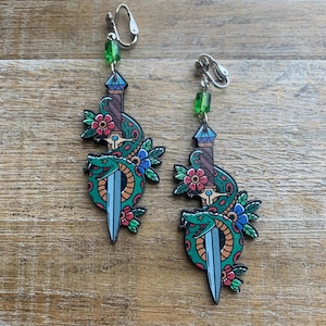 Pode incluir: Um par de brincos de pressão com um design de adaga com uma cobra verde, uma lâmina azul e flores vermelhas e azuis. Cada brinco tem uma conta verde no topo. Fundo de madeira.