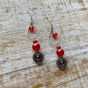 May include: A pair of dangle earrings featuring red glass beads and silver metal accents. The earrings have a teardrop-shaped wire frame with a smaller circle in the centre.