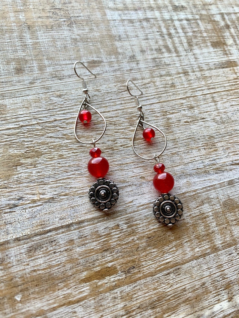 Op de afbeelding: Een paar oorhangers met rode glazen kralen en zilveren metalen accenten. De oorhangers hebben een traanvormig draadframe met een kleinere cirkel in het midden.