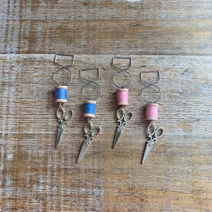 May include: Four miniature spools of thread with scissors attached. The spools are made of wood and have a blue, pink, pink, and pink thread wound around them. The scissors are silver and have a decorative design.