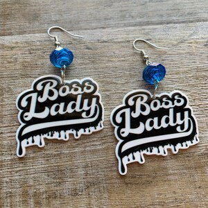 May include: A pair of earrings with a black and white "Boss Lady" design and blue crystal beads. The design has a dripping effect.