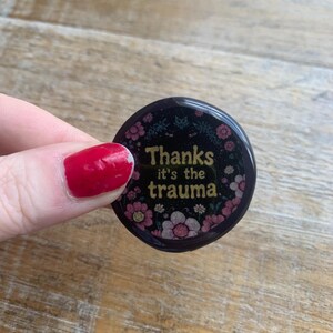 Könnte beinhalten: Ein runder, schwarzer Button mit dem Text "Thanks it's the trauma" in goldener Schrift. Der Button ist mit rosa und weißen Blumen verziert. Der Button wird von einer Person mit rotem Nagellack gehalten.