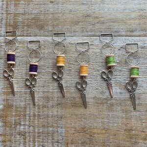 May include: Six sewing-themed charms with silver-colored scissors and thread spools in various colors. Each charm has a silver-colored metal loop and a wooden spool base. The thread colors include purple, yellow, and green.