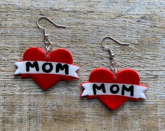 May include: A pair of red heart-shaped earrings with a white banner that says "MOM" in black letters. The earrings are on a wooden surface.