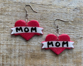 May include: A pair of red heart-shaped earrings with a white banner that says "MOM" on each earring. The earrings have silver hooks.