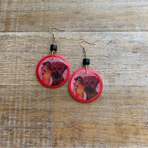 May include: A pair of dangle earrings featuring red circular pendants with a printed image of two figures. The earrings have gold-colored hooks and black square beads. The background is a wooden surface.