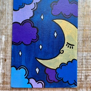 May include: A hand-painted canvas featuring a crescent moon with a sleeping face, surrounded by purple, blue, and light blue clouds against a dark blue background. The moon is yellow with a black outline and the clouds have black outlines. There are white diamond-shaped stars scattered across the canvas.
