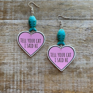 Puede incluir: Un par de pendientes en forma de corazón rosa con el texto "Tell your cat I said hi" y un dije de pez turquesa colgando de cada pendiente.