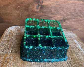 May include: A square, multi-compartment resin organizer in shades of teal and green. The organizer is decorated with glitter, giving it a sparkling appearance. The compartments are divided by a grid pattern.