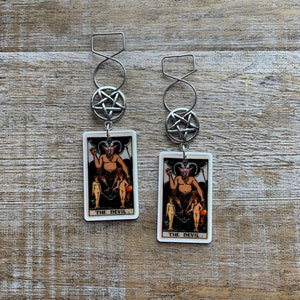 May include: A pair of silver earrings with a pentagram charm and a tarot card charm featuring the Devil card. The card is white with a vintage design and the text "THE DEVIL."