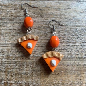May include: A pair of dangle earrings featuring a slice of pumpkin pie with a white dollop of whipped cream and an orange bead. The earrings are made of polymer clay and have silver hooks.
