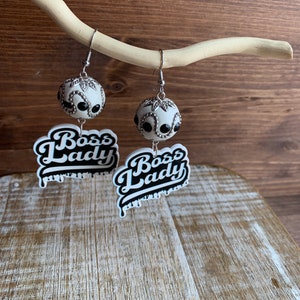 May include: A pair of dangle earrings with a white and black "Boss Lady" design. The earrings feature a white ball with black accents and silver beads.