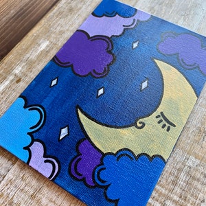 May include: A hand-painted canvas with a crescent moon and clouds in shades of blue, purple, and yellow. The moon has a face with closed eyes and a small smile.