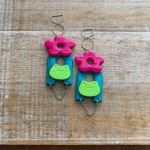 May include: A pair of turquoise and pink dangle earrings with a green frog charm and a flower charm. The earrings are made of polymer clay and have a silver chain.