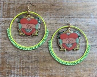 May include: A pair of large hoop earrings with a green and yellow beaded border. The earrings feature a heart-shaped charm with a hand-holding design.