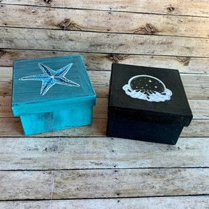 May include: Two small wooden boxes with painted designs. The box on the left is teal blue with a white starfish. The box on the right is black with a white image of a hand holding a crystal ball with stars.