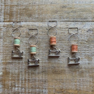 May include: Four silver sewing machine charms with spools of thread in green, orange, and peach colors. The charms are hanging from silver wire loops.