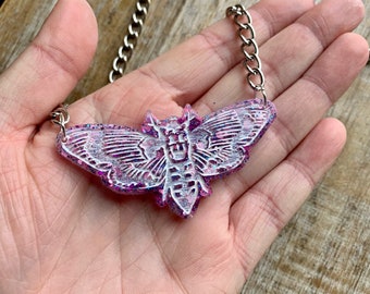 May include: A silver chain necklace with a pendant shaped like a moth. The moth pendant is white with intricate details and a glittery pink and purple background. The necklace is held in a hand.