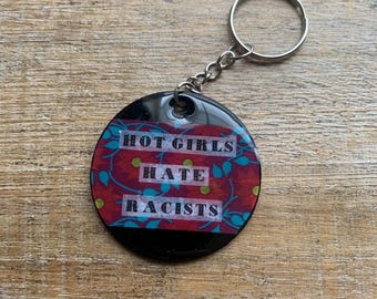 Könnte beinhalten: Runder, schwarzer Schlüsselanhänger mit dem weißen Text "HOT GIRLS HATE RACISTS". Der Schlüsselanhänger hat ein rotes und blaues Blumendesign. Er ist an einem silbernen Metallring befestigt.