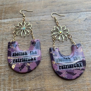May include: A pair of dangle earrings with a floral design and the text "abolish the patriarchy." The earrings feature a gold-colored flower and chain, with a purple and gold marbled design.