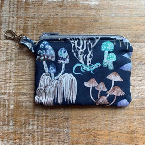 May include: A small, dark blue zippered pouch with a colorful mushroom and lizard print. The pouch has a silver zipper and a silver clip.