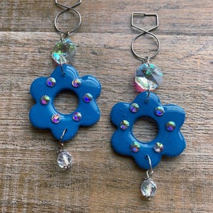 May include: A pair of blue flower-shaped earrings with iridescent rhinestones and a clear crystal bead dangle. The earrings have silver-colored metal findings and a clear crystal accent above the flower.